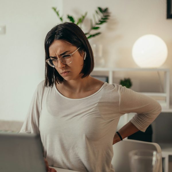 A person with glasses sits in front of a laptop, clutching their lower back with a pained expression. The scene highlights the struggle of chronic pain management in everyday life.