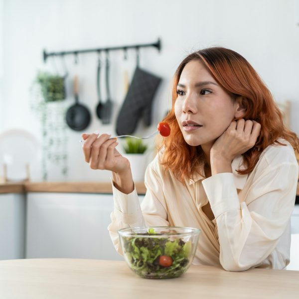 A person with auburn hair sits at a table, holding a fork with a cherry tomato, contemplating the bowl of salad in front of them. The kitchen background is brightly lit, creating an inviting atmosphere as they focus on nourishing both body and mental health.