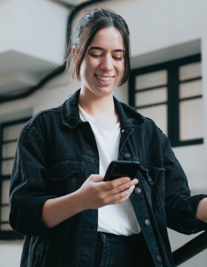 A person in a black jacket is smiling while looking at their phone, standing in front of a background with windows.