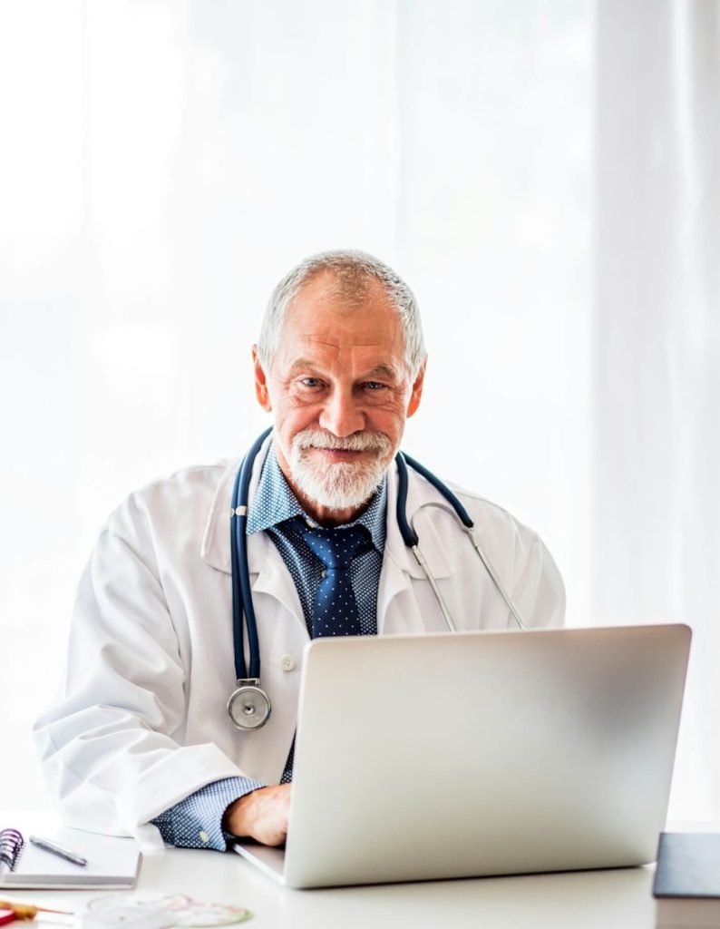 An elderly doctor with a stethoscope around his neck is smiling and sitting at a desk with a laptop.