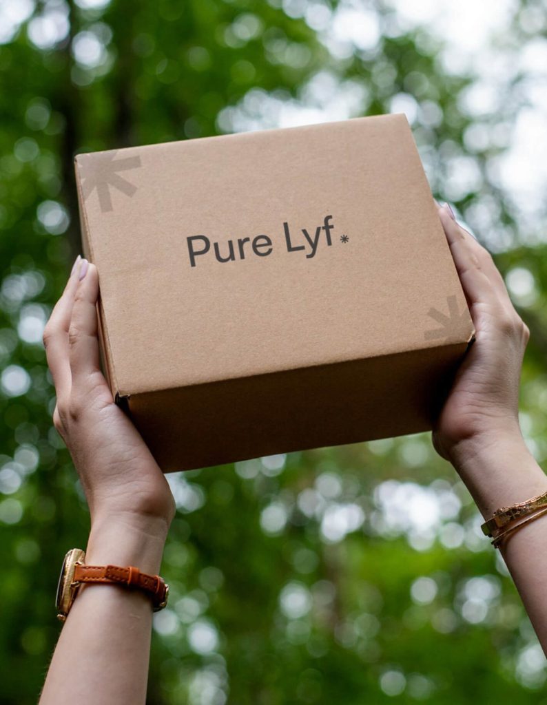 A person holds up a cardboard box labeled "Pure Lyf" against a background of green foliage.