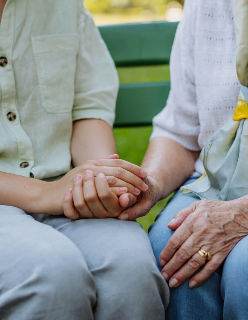 Two people sitting on a bench hold hands. One appears older, wearing a gold ring, and the other is younger. They're casually dressed in light-colored shirts and pants.
