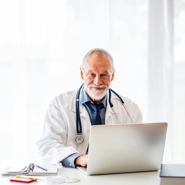 An older man in a white coat and stethoscope sits at a desk with a laptop, smiling at the camera.