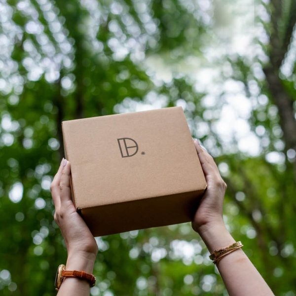 Hands holding a brown box with a logo against a background of green trees.